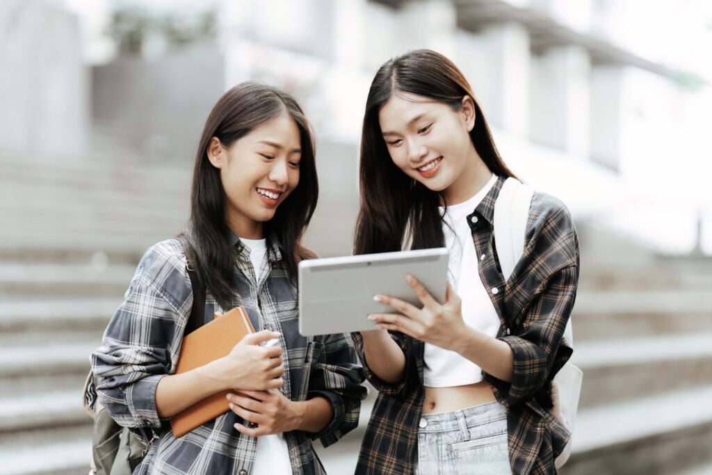 Two young Asian students smiling and studying together using a digital tablet outdoors, preparing for IELTS One Skill Boost.