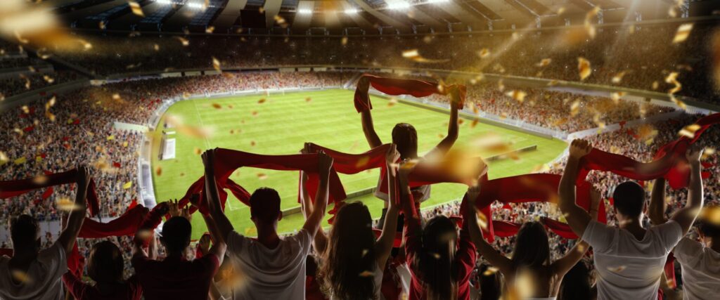 Fans celebrating in a football stadium with red scarves, representing fitness, professional sport, role models, and IELTS Sports and Leisure 2025.