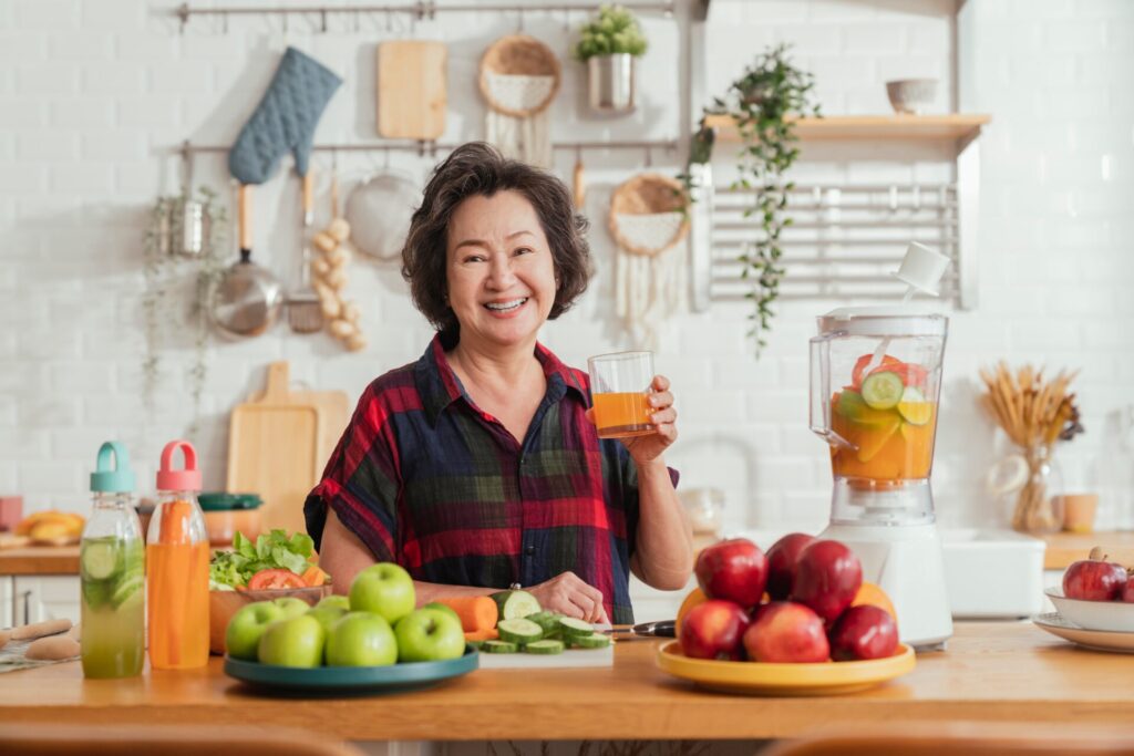 Smiling woman in kitchen holding fresh juice with apples, cucumber, and blender for IELTS Food and Lifestyle 2025 blog