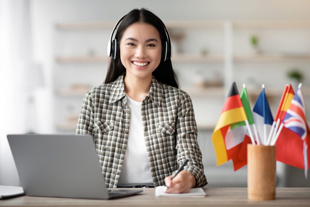 Smiling Asian student wearing headphones taking online English lesson at ILC Hua Hin, studying Conversation English with international flags in the background.